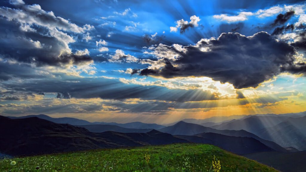 Ein Bild von Bergen, einer Wiese im Vordergrund und einem schönen, wolkigen Himmel, durch den Sonnenstrahlen kommen