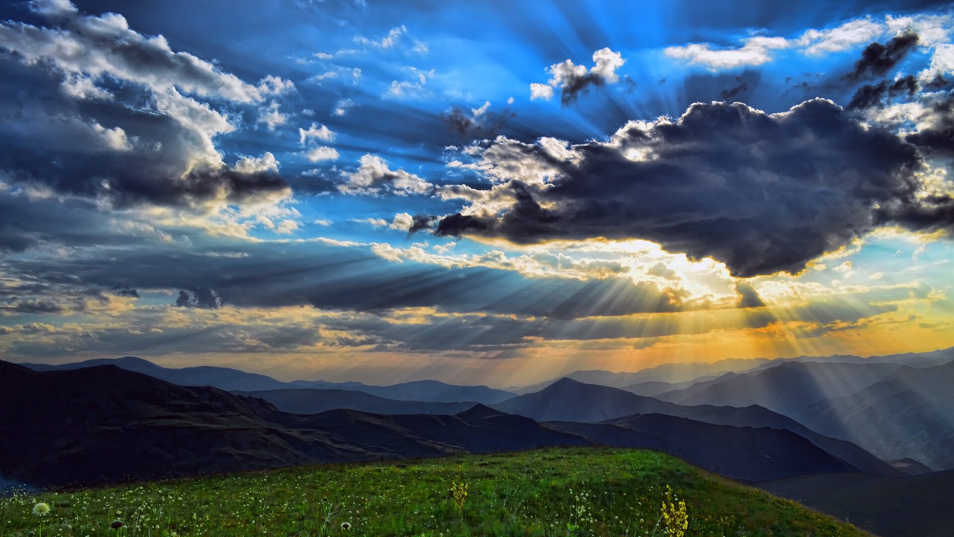 Ein Bild von Bergen, einer Wiese im Vordergrund und einem schönen, wolkigen Himmel, durch den Sonnenstrahlen kommen